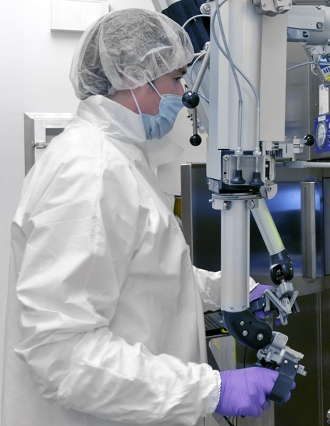 une photo d'un technicien de laboratoire en pleine préparation d'isopesticides, vêtu d'une blouse blanche et d'un bonnet à cheveux, derrière la vitre de sa machine